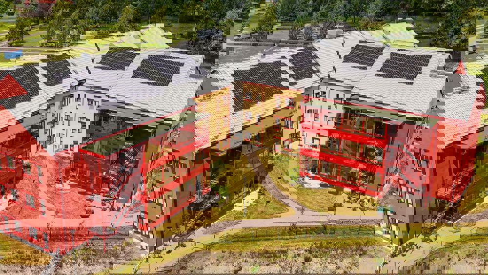 Röd och gul byggnad i L-form omgiven av grönska - Lövsta vård- och omsorgsboende